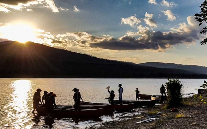 a group of outward bound students stand in shallow water beside their canoes as the sun is setting